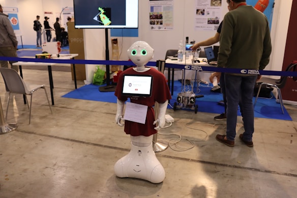 A humanoid robot with green-lit eyes and a screen on its chest displaying a logo stands in an exhibition space. It is surrounded by various pieces of equipment and people interacting with different technology displays. The setting appears to be industrial with smooth flooring, tables, chairs, and informational posters on the wall.