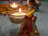 A beautifully decorated small figurine is holding a lit tealight candle. Around the figure, there are more delicate decorations and intricate patterns, including marigold flowers set on a colorful background. The ambiance is festive, warm, and inviting.