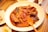 A large white plate contains a dish of spicy, red-colored fish stew, garnished with sesame seeds and vegetables. An orange plastic spoon lies on the side of the plate. The table also has small side dishes with cabbage salad and some type of condiment.