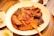 A large white plate contains a dish of spicy, red-colored fish stew, garnished with sesame seeds and vegetables. An orange plastic spoon lies on the side of the plate. The table also has small side dishes with cabbage salad and some type of condiment.