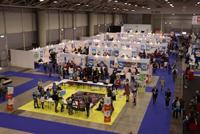 A busy exhibition hall with numerous booths lined up in rows. People are gathered around tables and booths, engaging in activities and conversations. The floors are covered with vibrant blue and yellow carpeting, creating distinct areas for different displays. Overhead lights provide bright illumination in the spacious indoor setting.