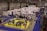A busy exhibition hall with numerous booths lined up in rows. People are gathered around tables and booths, engaging in activities and conversations. The floors are covered with vibrant blue and yellow carpeting, creating distinct areas for different displays. Overhead lights provide bright illumination in the spacious indoor setting.