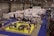 A busy exhibition hall with numerous booths lined up in rows. People are gathered around tables and booths, engaging in activities and conversations. The floors are covered with vibrant blue and yellow carpeting, creating distinct areas for different displays. Overhead lights provide bright illumination in the spacious indoor setting.