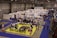 A busy exhibition hall with numerous booths lined up in rows. People are gathered around tables and booths, engaging in activities and conversations. The floors are covered with vibrant blue and yellow carpeting, creating distinct areas for different displays. Overhead lights provide bright illumination in the spacious indoor setting.
