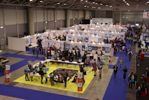 A busy exhibition hall with numerous booths lined up in rows. People are gathered around tables and booths, engaging in activities and conversations. The floors are covered with vibrant blue and yellow carpeting, creating distinct areas for different displays. Overhead lights provide bright illumination in the spacious indoor setting.
