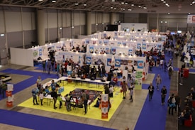 A busy exhibition hall with numerous booths lined up in rows. People are gathered around tables and booths, engaging in activities and conversations. The floors are covered with vibrant blue and yellow carpeting, creating distinct areas for different displays. Overhead lights provide bright illumination in the spacious indoor setting.