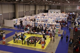 A busy exhibition hall with numerous booths lined up in rows. People are gathered around tables and booths, engaging in activities and conversations. The floors are covered with vibrant blue and yellow carpeting, creating distinct areas for different displays. Overhead lights provide bright illumination in the spacious indoor setting.
