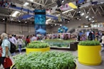 An indoor exhibition or trade show filled with people walking around various displays. Large planters filled with green plants are prominently situated in the foreground. There is a tall digital display screen showing advertisements and graphics, with bright overhead lighting fixtures. Hanging spheres resembling Earth globes and a banner reading 'Blue Loop' are visible. The setting is spacious and modern with a busy and vibrant atmosphere.