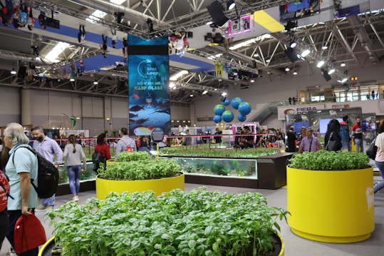 An indoor exhibition or trade show filled with people walking around various displays. Large planters filled with green plants are prominently situated in the foreground. There is a tall digital display screen showing advertisements and graphics, with bright overhead lighting fixtures. Hanging spheres resembling Earth globes and a banner reading 