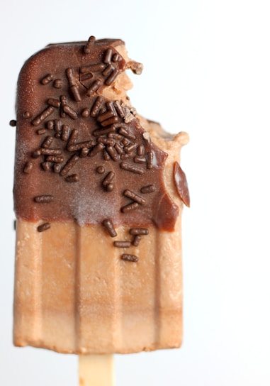 A popsicle with a chocolate coating and chocolate sprinkles, partially eaten with a bite taken out of the top.