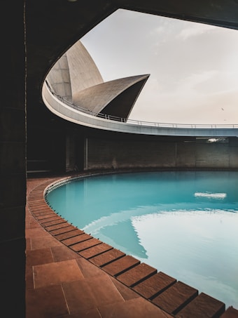 A modern architectural structure featuring smooth, curved concrete walls and a large, shallow pool of water. The setting includes a tiled walkway around the water, creating an elegant contrast. Natural light filters from above, highlighting the geometric lines and textures.