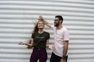 A man and a woman stand in front of a textured white wall. The woman has long red hair and is wearing a green T-shirt with purple pants, smiling with her eyes closed, while the man in a light pink T-shirt is laughing and playfully holding her hair. Both appear relaxed and joyful.
