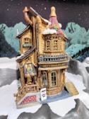 A miniature model of a chocolate shop is set in a snowy, mountainous landscape. The shop features a wooden facade with detailed windows and a sign that reads 'Lucy's Chocolate Shop'. Snow covers parts of the roof and surrounding ground. A small sign outside advertises 'Free samples today'. The background displays snow-capped mountains under a starry sky.