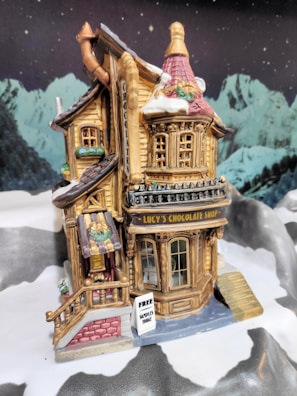 A miniature model of a chocolate shop is set in a snowy, mountainous landscape. The shop features a wooden facade with detailed windows and a sign that reads 'Lucy's Chocolate Shop'. Snow covers parts of the roof and surrounding ground. A small sign outside advertises 'Free samples today'. The background displays snow-capped mountains under a starry sky.