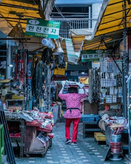 A narrow alley in a market is lined with stalls selling various electronics and hardware items. The ceiling is partially covered with yellow and green fabric awnings. Signs with Korean writing hang above the stalls. A person in a pink patterned outfit and red pants is carrying a load above their head while walking through the alley, creating a bustling atmosphere.