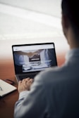 A person sits at a wooden table, using a laptop. The screen displays a website with images of a handshake and some text in Italian. Nearby, a notebook and a pen rest on the table.