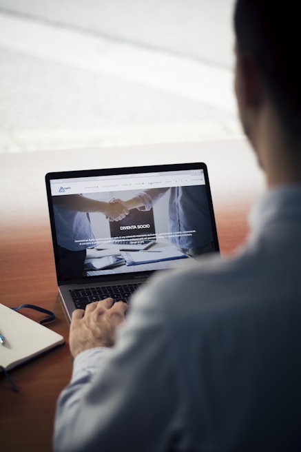 A person sits at a wooden table, using a laptop. The screen displays a website with images of a handshake and some text in Italian. Nearby, a notebook and a pen rest on the table.