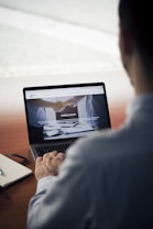 A person sits at a wooden table, using a laptop. The screen displays a website with images of a handshake and some text in Italian. Nearby, a notebook and a pen rest on the table.