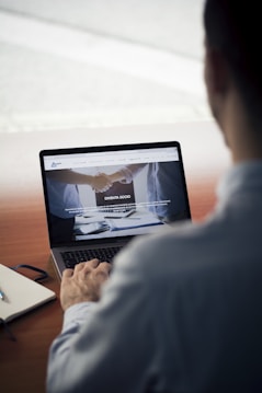 A person sits at a wooden table, using a laptop. The screen displays a website with images of a handshake and some text in Italian. Nearby, a notebook and a pen rest on the table.