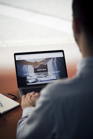 A person sits at a wooden table, using a laptop. The screen displays a website with images of a handshake and some text in Italian. Nearby, a notebook and a pen rest on the table.