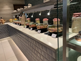 A buffet setup with multiple pots of food placed on a counter. The pots are covered with clear lids, allowing a view of the dishes inside. Behind the pots, there are jars with red and white checkered cloth covers, possibly containing grains or spices. The countertop is sleek and white, contrasted by the dark tiled backsplash. Overhead lights highlight the food items.