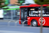 A bright red city bus with advertising for free Wi-Fi powered by a telecommunications company drives along a street. The bus appears to be in motion, and partially obscured by a tree. The background is slightly blurred, indicating speed. The word 'CROWN' is displayed on the bus’s digital sign.