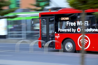 A bright red city bus with advertising for free Wi-Fi powered by a telecommunications company drives along a street. The bus appears to be in motion, and partially obscured by a tree. The background is slightly blurred, indicating speed. The word 'CROWN' is displayed on the bus’s digital sign.