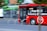 A bright red city bus with advertising for free Wi-Fi powered by a telecommunications company drives along a street. The bus appears to be in motion, and partially obscured by a tree. The background is slightly blurred, indicating speed. The word 'CROWN' is displayed on the bus’s digital sign.