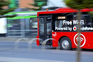 A bright red city bus with advertising for free Wi-Fi powered by a telecommunications company drives along a street. The bus appears to be in motion, and partially obscured by a tree. The background is slightly blurred, indicating speed. The word 'CROWN' is displayed on the bus’s digital sign.