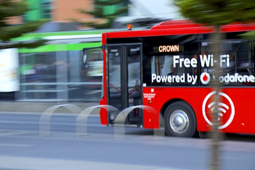 A bright red city bus with advertising for free Wi-Fi powered by a telecommunications company drives along a street. The bus appears to be in motion, and partially obscured by a tree. The background is slightly blurred, indicating speed. The word 'CROWN' is displayed on the bus&rsquo;s digital sign.