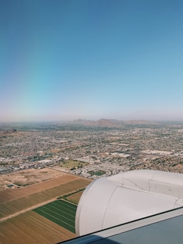 An airplane's engine is visible as the plane flies over a sprawling landscape below. The terrain includes a patchwork of agricultural fields, urban areas, and open spaces. Distant mountains rise in the background under a clear blue sky.