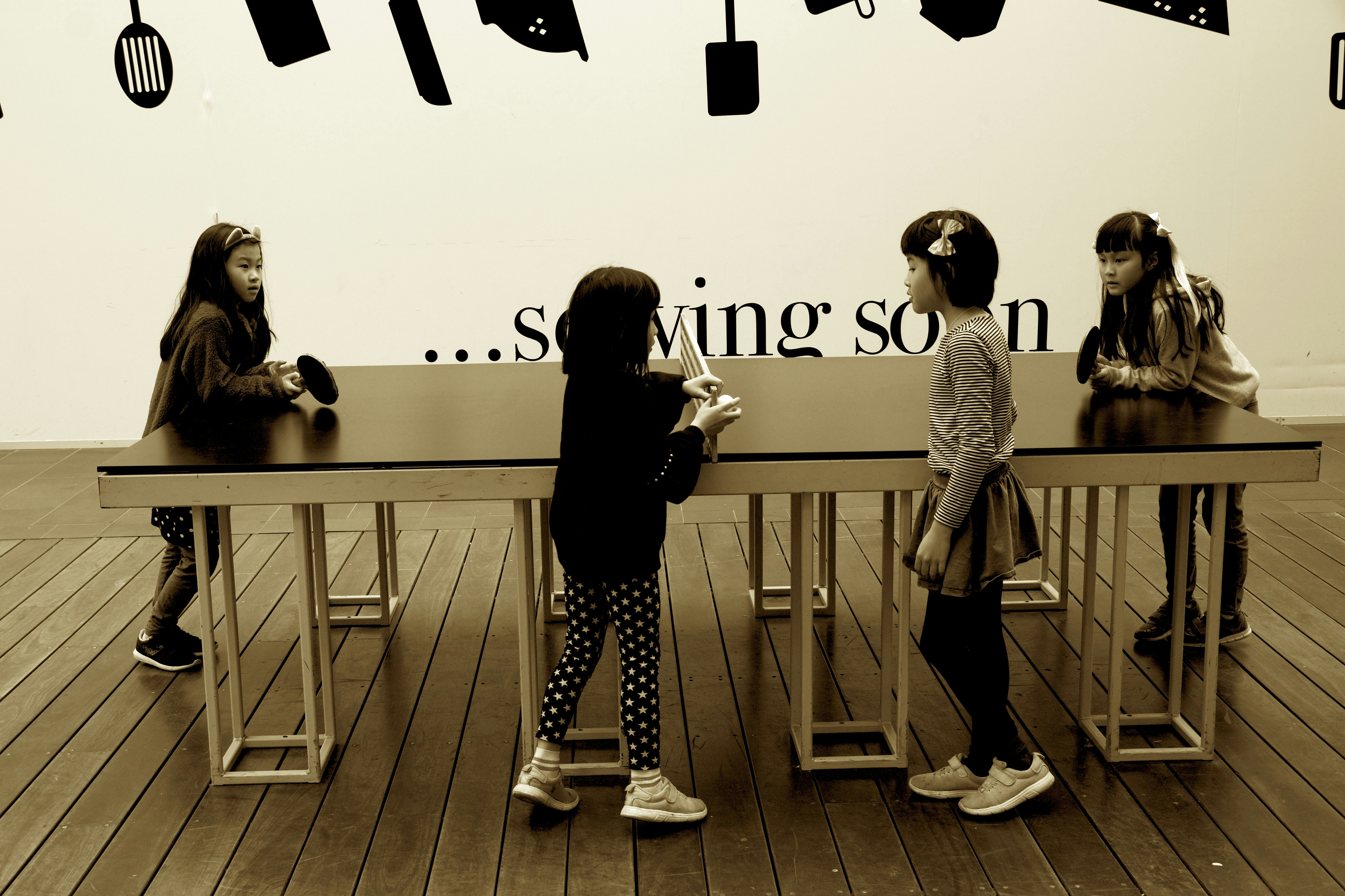 Four young children are interacting around a long, rectangular table with a smooth surface. The setting includes a wooden floor and a wall decorated with silhouettes of kitchen utensils. Above the table, the text on the wall partially reads '... sowing soon'.