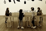 Four young children are interacting around a long, rectangular table with a smooth surface. The setting includes a wooden floor and a wall decorated with silhouettes of kitchen utensils. Above the table, the text on the wall partially reads '... sowing soon'.
