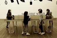 Four young children are interacting around a long, rectangular table with a smooth surface. The setting includes a wooden floor and a wall decorated with silhouettes of kitchen utensils. Above the table, the text on the wall partially reads '... sowing soon'.