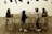 Four young children are interacting around a long, rectangular table with a smooth surface. The setting includes a wooden floor and a wall decorated with silhouettes of kitchen utensils. Above the table, the text on the wall partially reads '... sowing soon'.