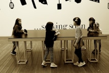 Four young children are interacting around a long, rectangular table with a smooth surface. The setting includes a wooden floor and a wall decorated with silhouettes of kitchen utensils. Above the table, the text on the wall partially reads '... sowing soon'.