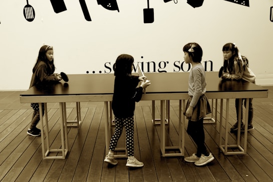 Four young children are interacting around a long, rectangular table with a smooth surface. The setting includes a wooden floor and a wall decorated with silhouettes of kitchen utensils. Above the table, the text on the wall partially reads '... sowing soon'.