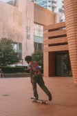 A person in a colorful Hawaiian shirt and camouflage pants is skateboarding on a wide, tiled plaza. Tall modern buildings and trees are visible in the background, lending an urban feel to the scene.