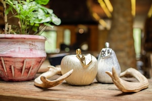A decorative setup featuring a potted plant with lush green leaves in a pinkish ceramic pot. Beside it, there are metallic decorative fruits shaped like an apple and a pear with shiny surfaces. Two antler pieces are artfully placed nearby, all arranged on a rustic wooden table.