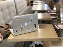A laptop decorated with various stickers is placed on a wooden table. Beside it, there's a small plate with a cookie. In the background, multiple tables and chairs are arranged in a neat row, giving a casual cafe or library vibe. A beige cushioned bench lines the left side of the image.