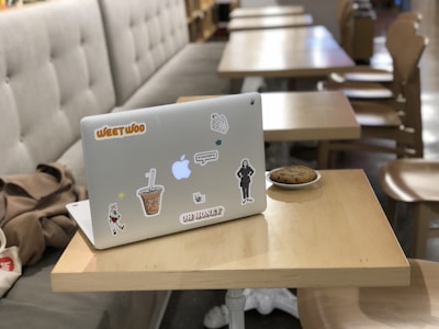 A laptop decorated with various stickers is placed on a wooden table. Beside it, there's a small plate with a cookie. In the background, multiple tables and chairs are arranged in a neat row, giving a casual cafe or library vibe. A beige cushioned bench lines the left side of the image.