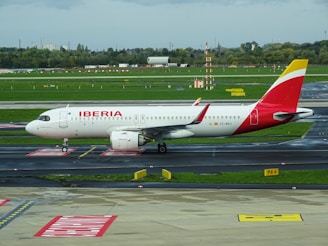 A passenger airplane with the Iberia livery is positioned on a taxiway at an airport. The aircraft is white with red and yellow accents and displays the Iberia logo prominently on the fuselage. The surroundings include a runway and green grass along with some airport signage and infrastructure in the background.