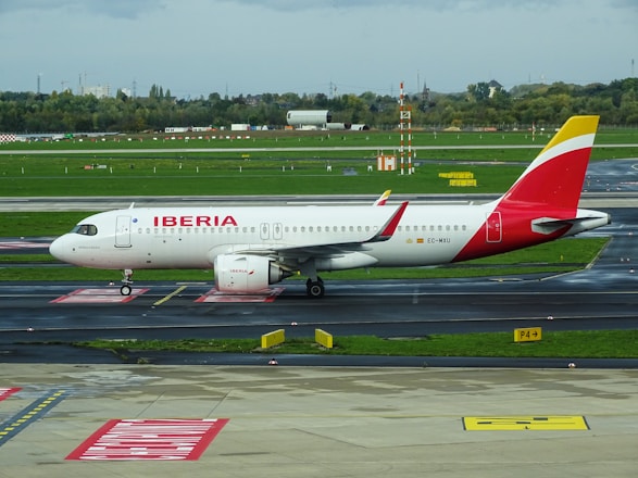 A passenger airplane with the Iberia livery is positioned on a taxiway at an airport. The aircraft is white with red and yellow accents and displays the Iberia logo prominently on the fuselage. The surroundings include a runway and green grass along with some airport signage and infrastructure in the background.