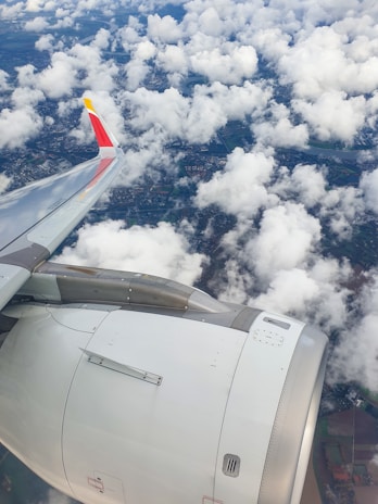 Aerial view of an airplane wing and engine against a backdrop of scattered clouds and the landscape below. The colors are vibrant, with the airplane featuring a sleek design and a distinct yellow, red, and white tail fin.