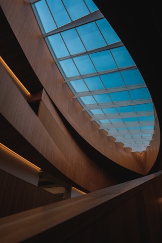 An interior architectural design featuring a large skylight with blue-tinted glass panels. The structure is characterized by smooth, curved wooden elements that create a modern and elegant aesthetic. The lighting is soft and focused, creating contrasting shadows and a warm ambiance.