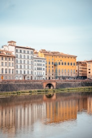 A row of colorful, classic European buildings line a waterfront. The buildings reflect in the calm water below, creating a mirrored effect. The architecture features large windows and ornate details, with colors ranging from pastel blue to vibrant yellow. A rustic stone bridge can be seen in the foreground.