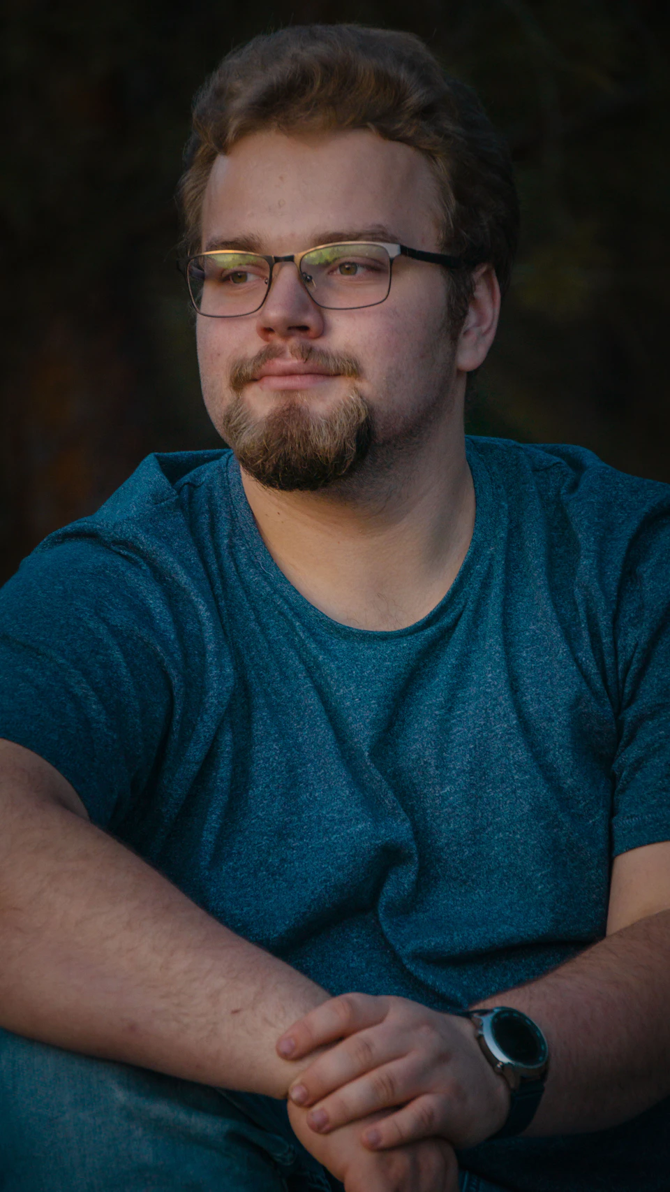 The image shows a young man with glasses sitting on a bench, dressed casually in a blue shirt and black pants. He is looking off to the side, possibly observing something or someone out of frame. The background appears to be a park setting, suggesting that he might be taking a break from his daily activities.
