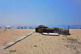 A sandy beach with a wooden walkway leading towards the shore. Multiple thatched and colorful umbrellas are set up near the water, where people are enjoying the beach. There is an old, graffiti-covered concrete structure partially buried in the sand.