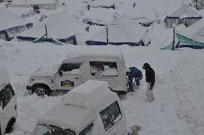 A snow-covered landscape with several vehicles partially buried under snow. Two individuals are working to clear snow from a white jeep labeled 'HIMALAYAN MOTOR SPORT'. Tents with snow-covered roofs are scattered in the background.