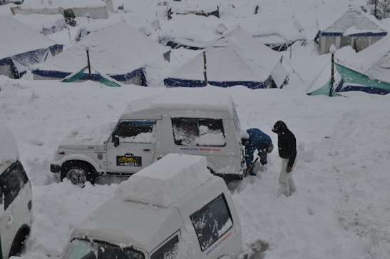 A snow-covered landscape with several vehicles partially buried under snow. Two individuals are working to clear snow from a white jeep labeled 'HIMALAYAN MOTOR SPORT'. Tents with snow-covered roofs are scattered in the background.
